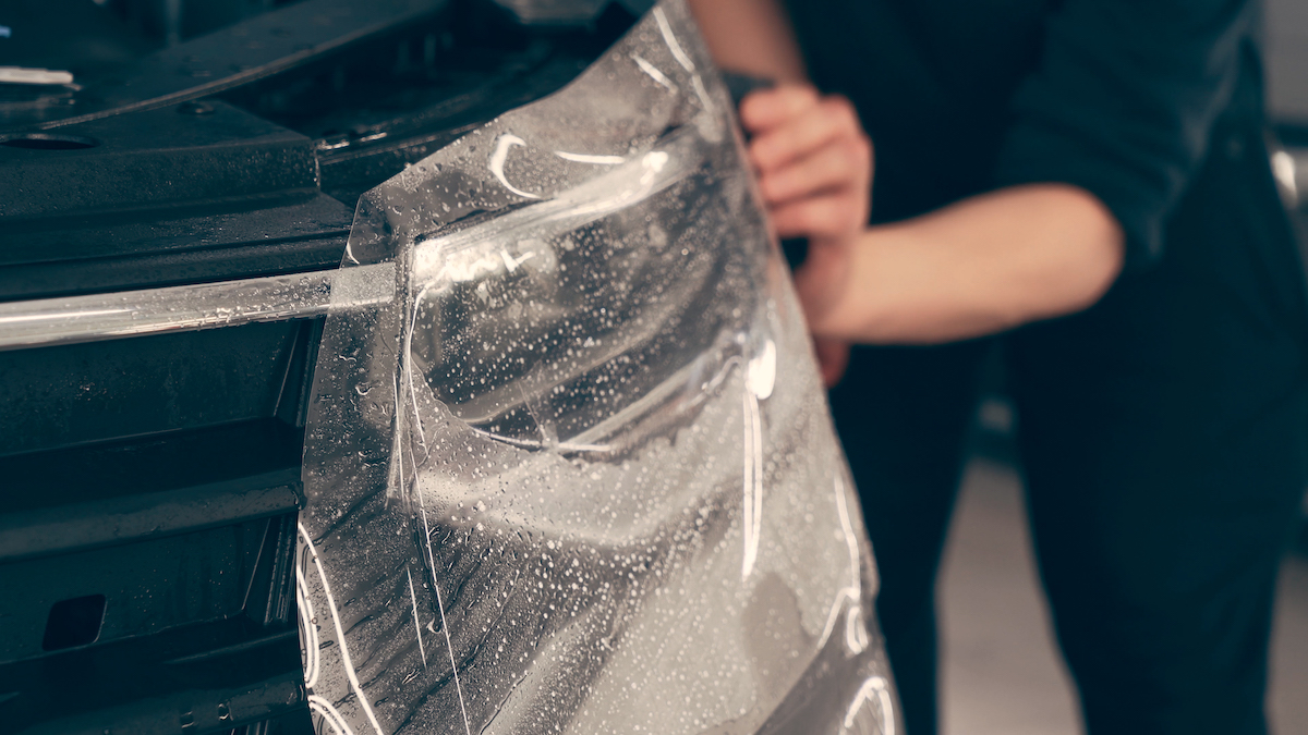 Detailer worker smoothes PPF or Paint Protection Film on Car Headlight with working tool. Polymer protective outer coating, close up.